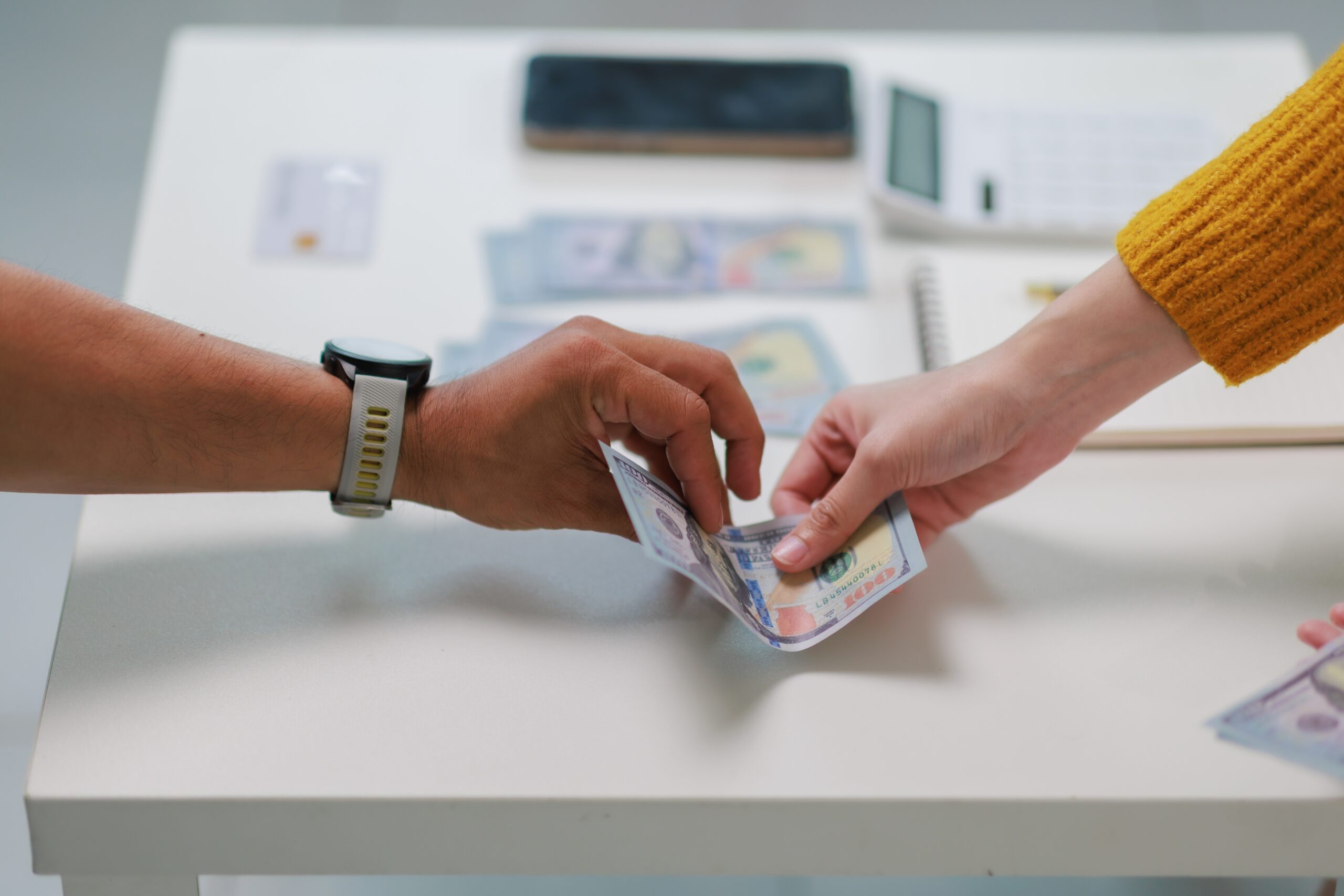 Two people exchanging hundred dollar bills across a desk with financial documents in the background.