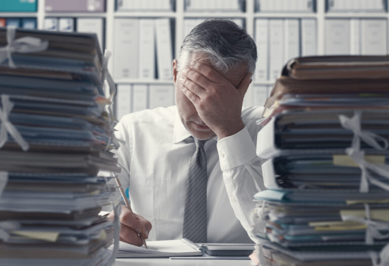 Overwhelmed office worker holding his forehead at a desk between tall stacks of paperwork.